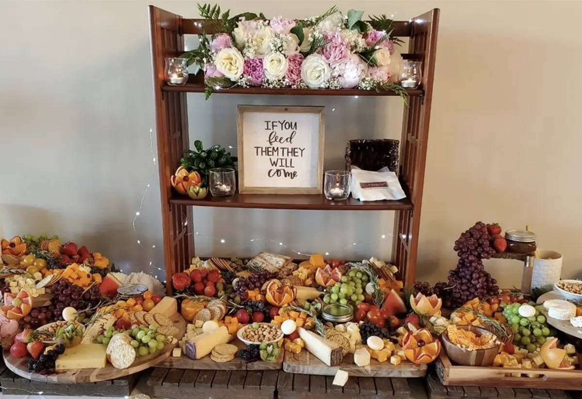 Close-up photograph perspective of a shelf containing small flower bouquet decorations, a picture frame "If you feed them they will come", and other assorted wedding objects while there are additionally wooden trays with food on top of them located nearby the shelf such as cheese, fruits, crackers, etc.