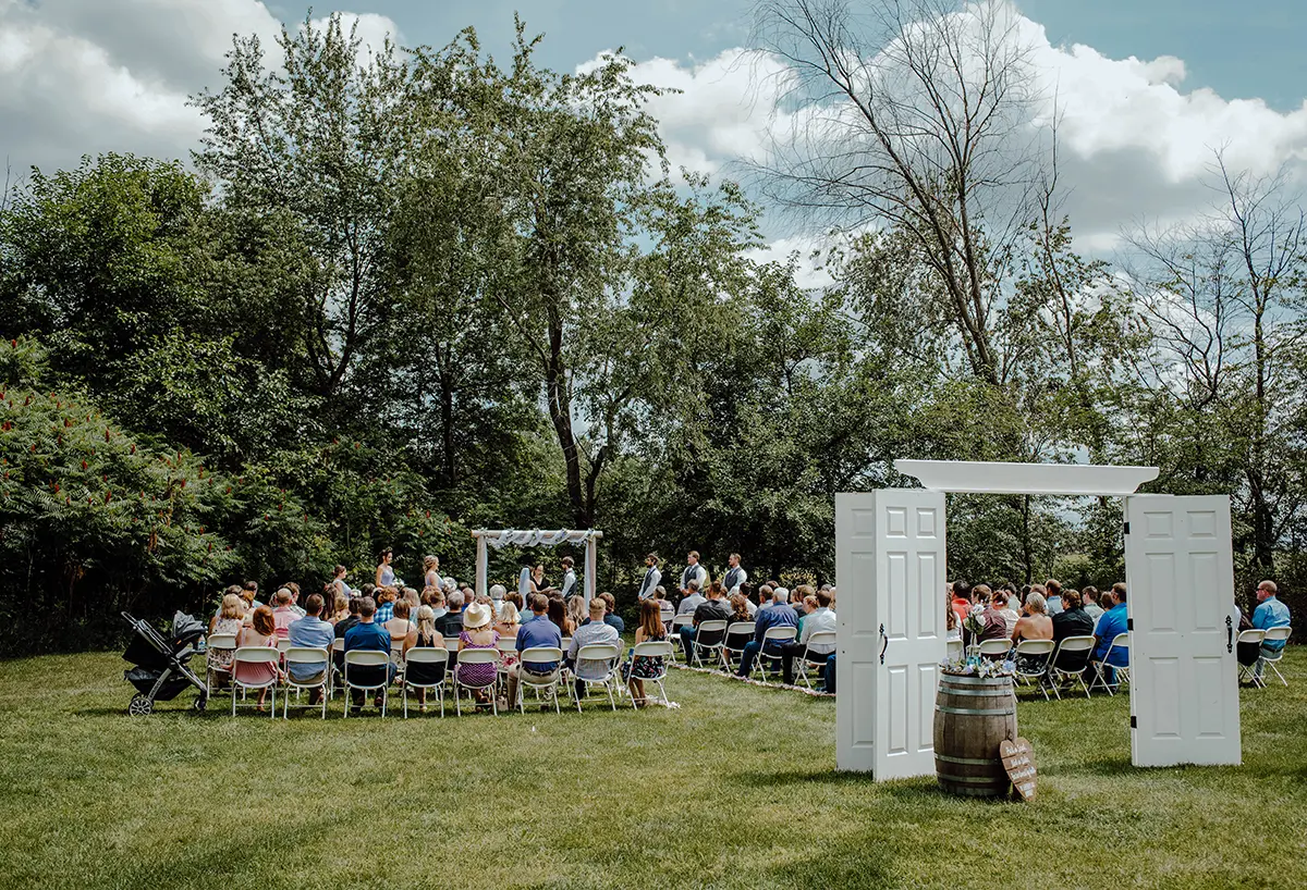 Wedding ceremony in session in a grassy lawn area with the bride and groom getting married as the audience glances upon them outside at the District 5 Schoolhouse venue