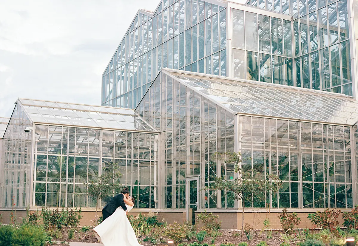 Bride and groom kiss each other at an angle downward as they are standing outside a glass building at Frederik Meijer Gardens & Sculpture Park