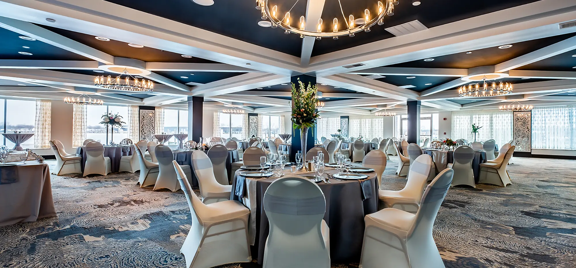Large room area with dark grey tables all around, dark tan chairs, glass cups, plates, forks, knifes, big circular glass lit chandeliers, and many other wedding assortment objects inside at the Holiday Inn Spring Lake-Grand Haven venue