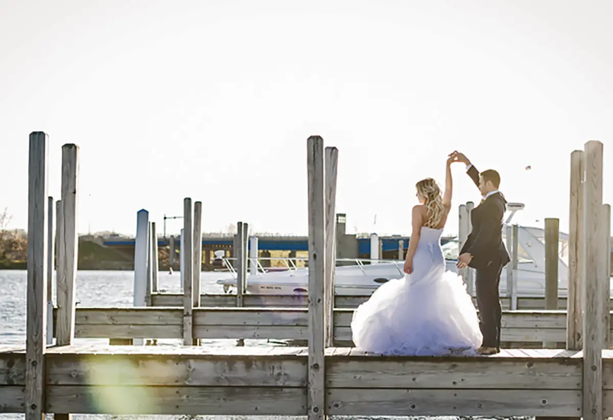 Outdoor landscape photograph of a bride and groom looking out into the water as the groom twirls/spins the bride in circles with his hands as they are standing on a dock area with a nearby small yacht in the background at the Holiday Inn Spring Lake-Grand Haven venue