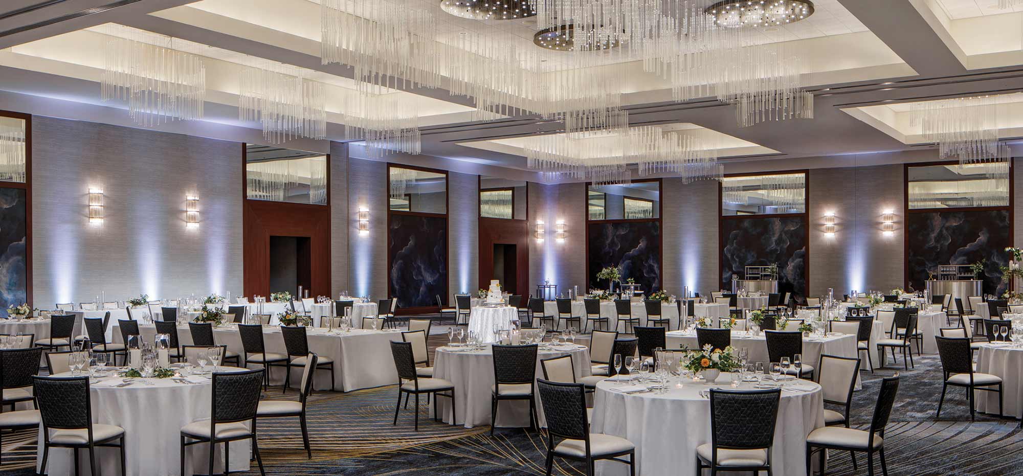 Large ballroom area with many white tables and black colored chairs all around with glass cups, forks, spoons, napkins, small flower bouquet decorations, and hanging floating crystal thin skinny chandeliers inside at the JW Marriott Grand Rapids venue