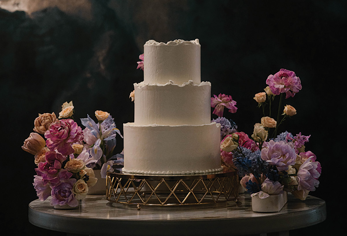 Close-up photograph perspective of a three layered decorative white cake and nearby surrounding flower bouquet decorations on a table inside the JW Marriott Grand Rapids venue