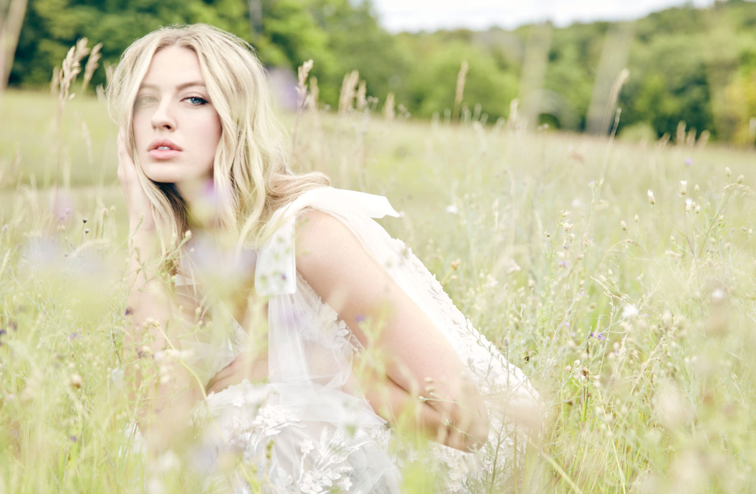 blonde haired female model wearing the Lillian West Verona beaded gown with detachable shoulder bows while sitting in a meadow