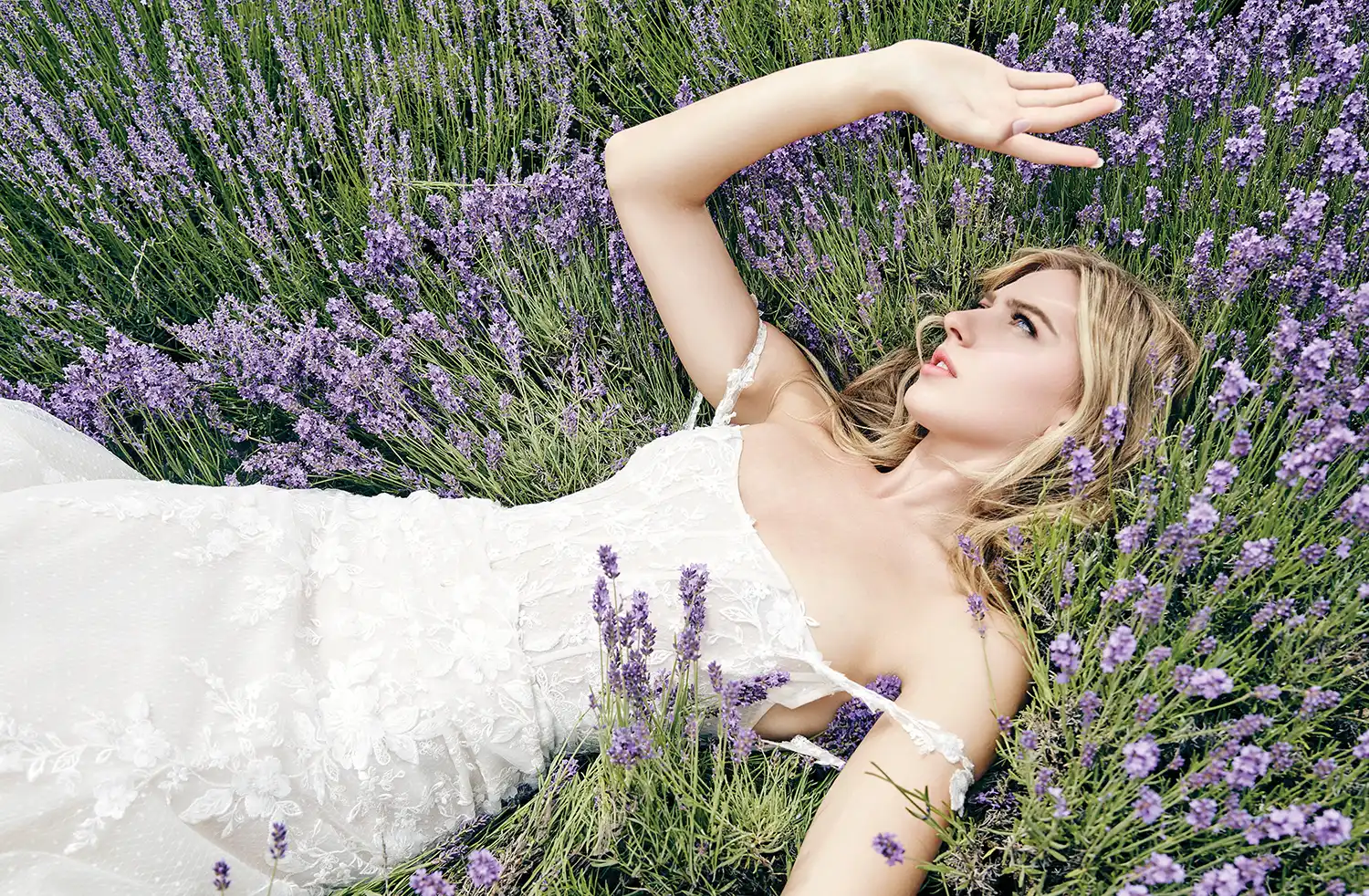 blonde haired female model laying in a meadow while wearing the Ines di Santo Marz lace applique corseted gown with draped off-the-shoulder sleeves