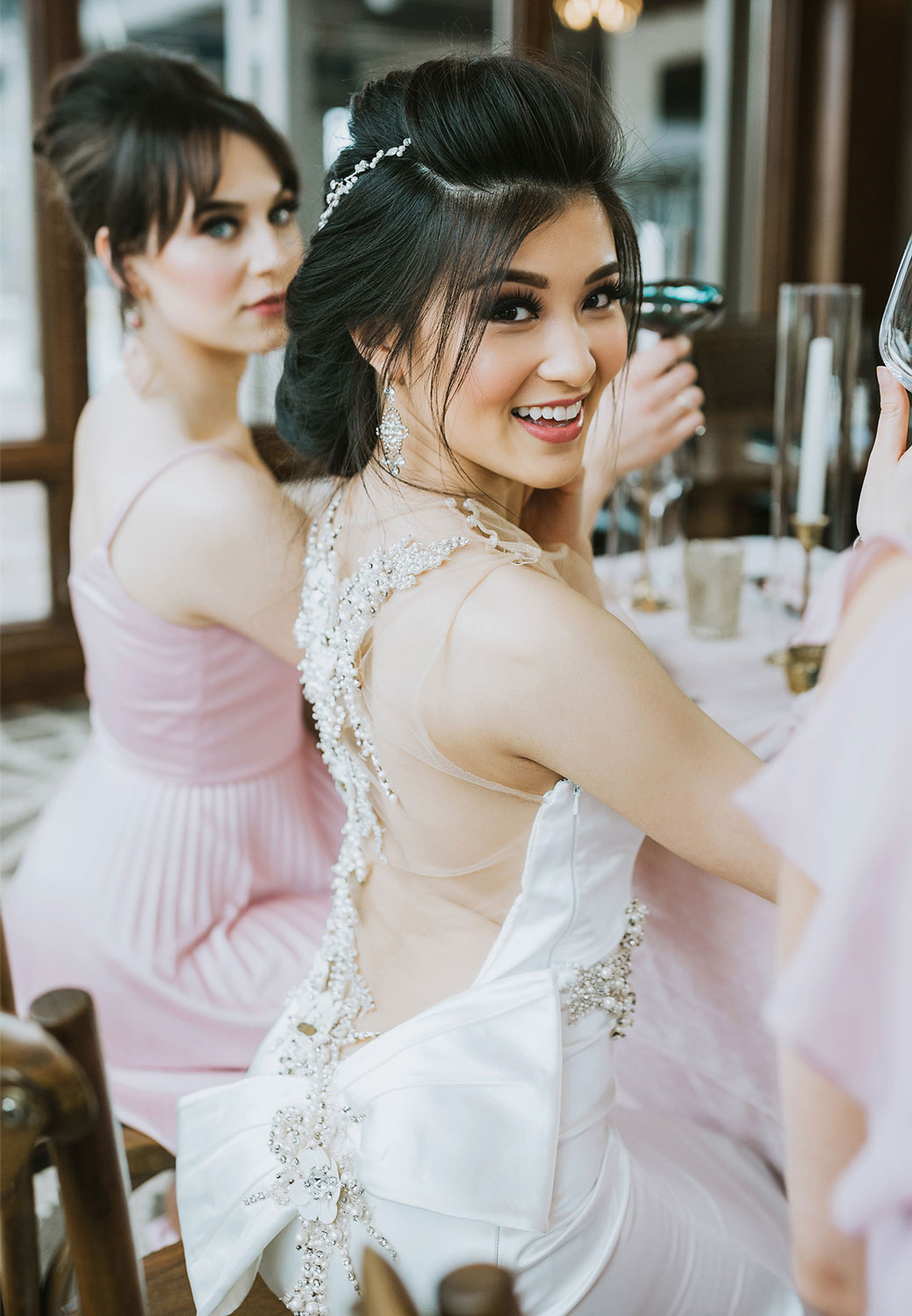 a bride looks over her shoulder at the camera while sitting at a table and raising a glass, a bridesmaid sits beside her slightly out of focus
