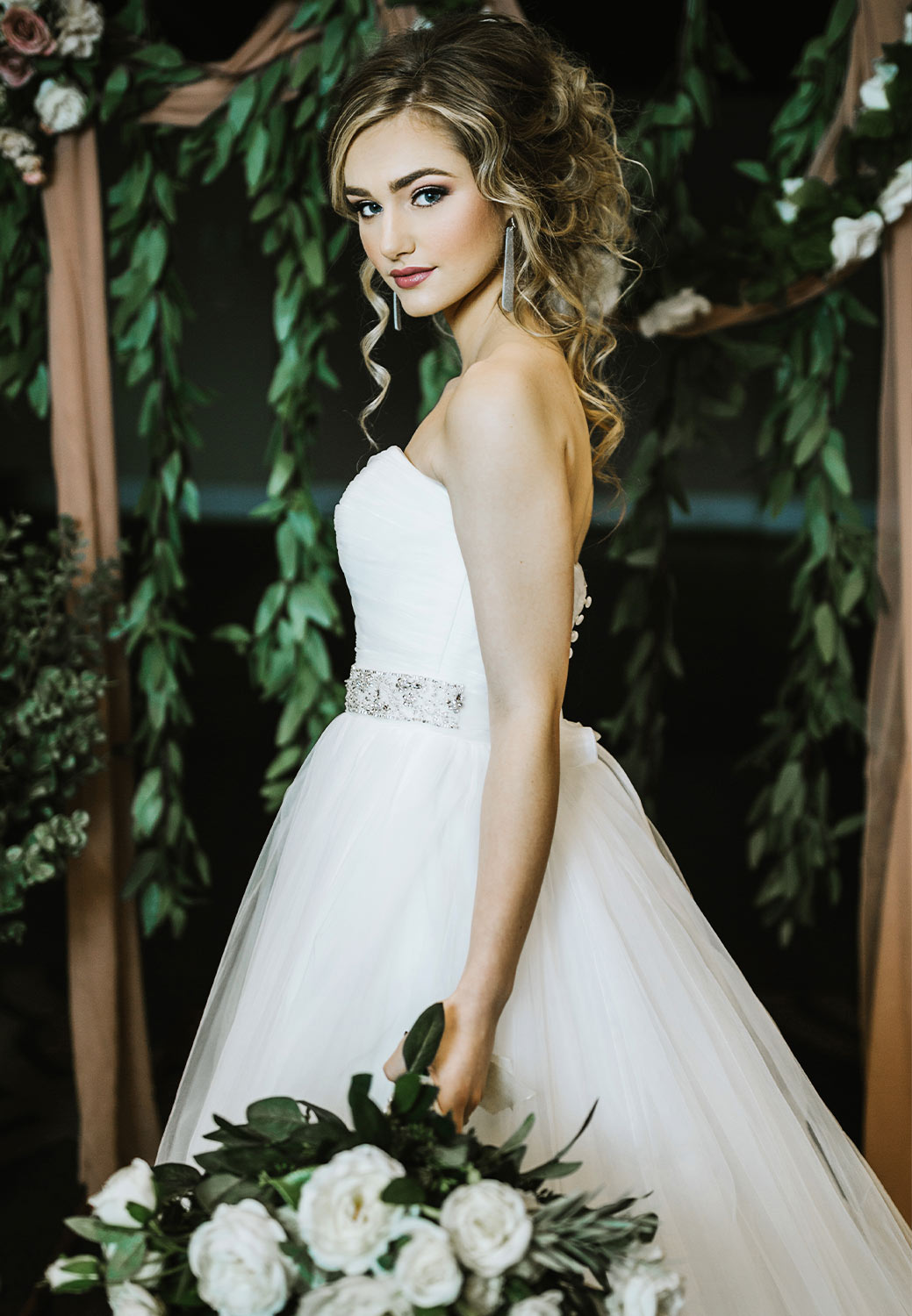 a young bride in a straight neckline gown poses for a wedding photo while holding a large bouquet and standing against a background of leaves and drapery
