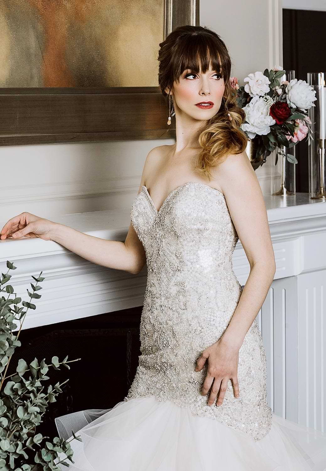 a bride in a sweetheart neckline mermaid wedding dress poses for a photo, standing beside a fireplace and looking away from the camera