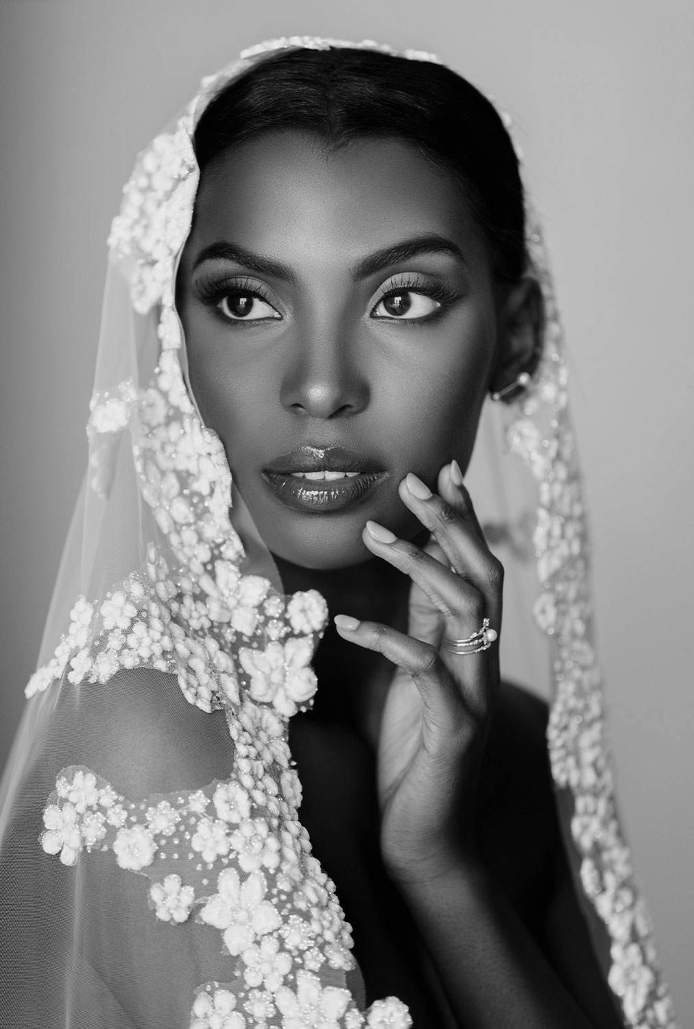 Close black and white portrait image of a woman wearing a Pronovias les Accessories Lucrezia mantilla style cathedral veil, a pear diamond ring and pearl earrings. She softly rests her manicured hand on her low cheek and looks beyond the camera.