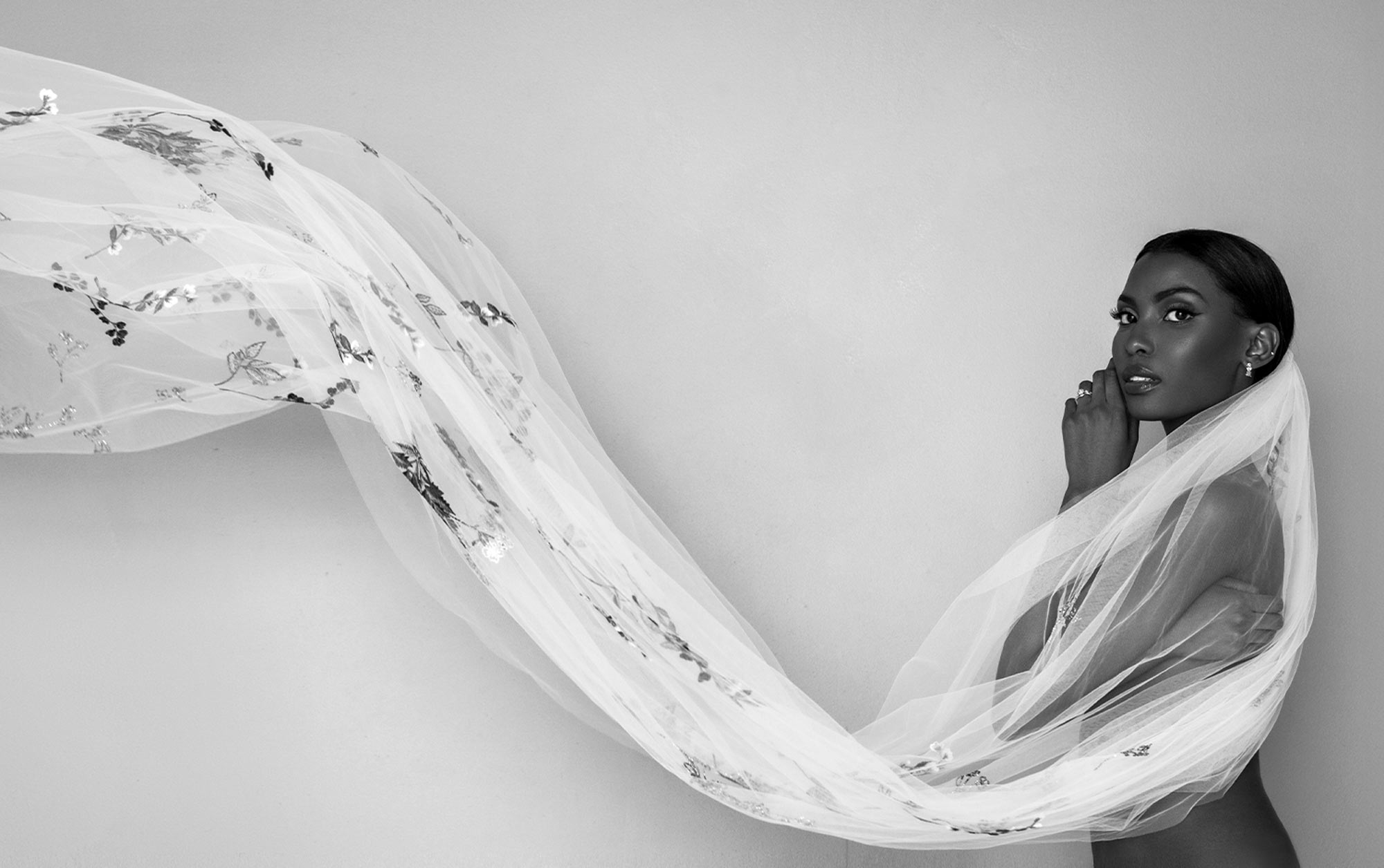 black and white wide view of a woman in a profile pose wearing a long flowing Theia Jasper floral embroidered veil that wafts out of left frame