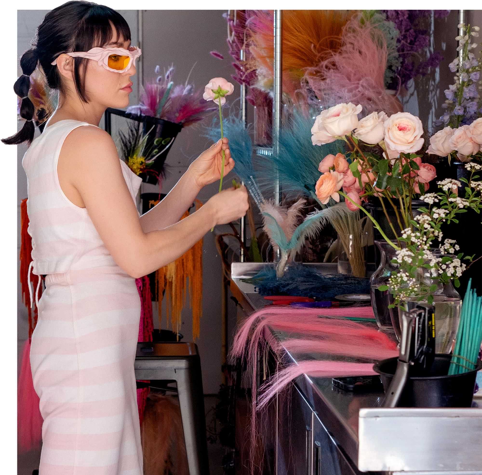 side view of Lindsay McWhorter, wearing a striped pink outfit and pink twisted rim glasses, stands at counter in a room full of colorful supplies while working on a pink rose arrangement
