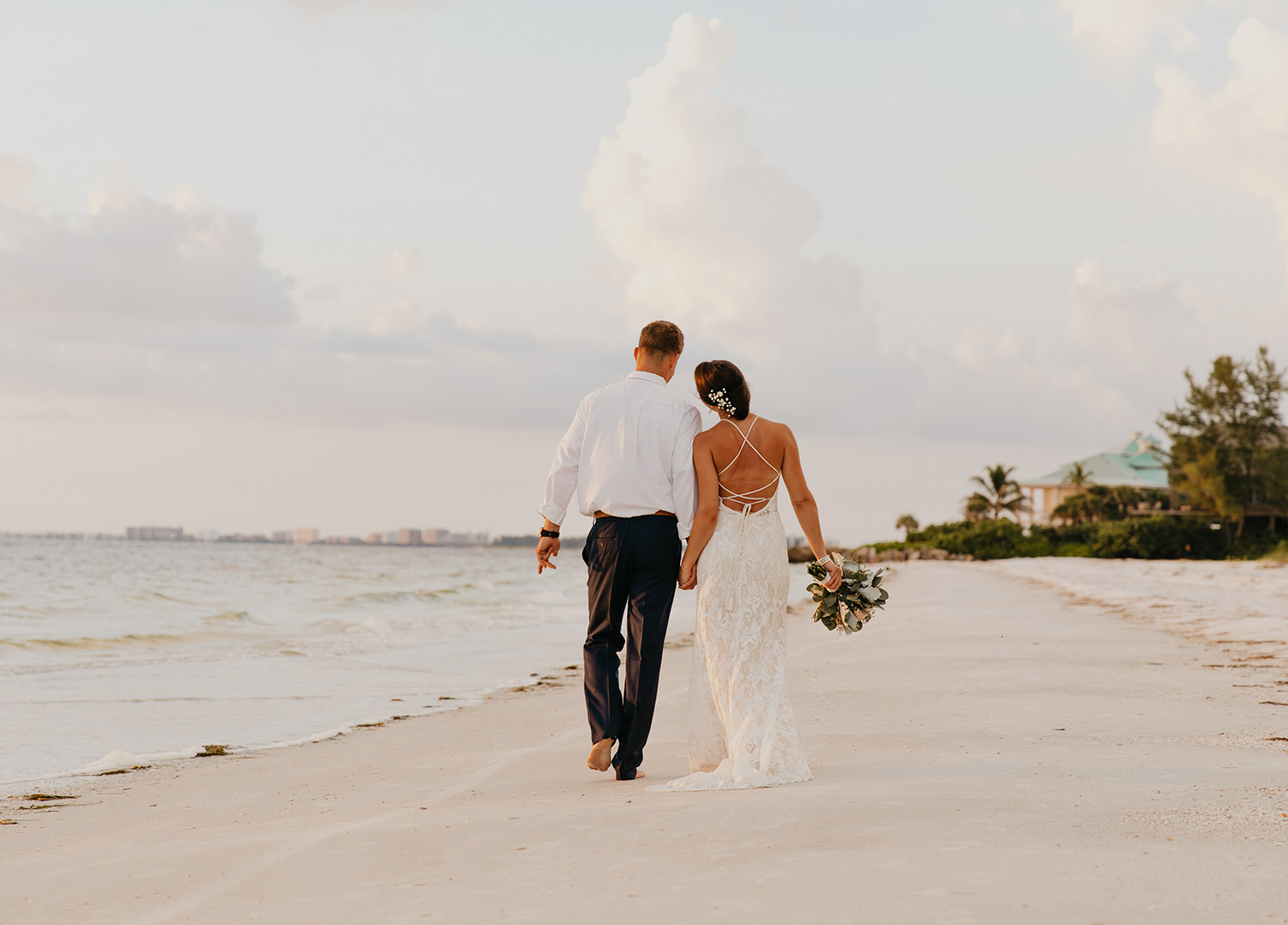 back view of a man in a white button up and black slacks and woman wearing a strappy back sheath wedding dress walking along a beach midday while holding hands