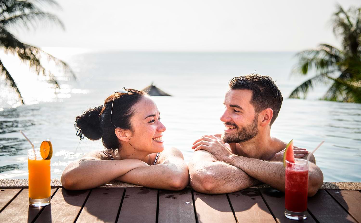 close up of a woman and man in water resting their arms on a small boardwalk where their drinks sit, they both look at each other smiling