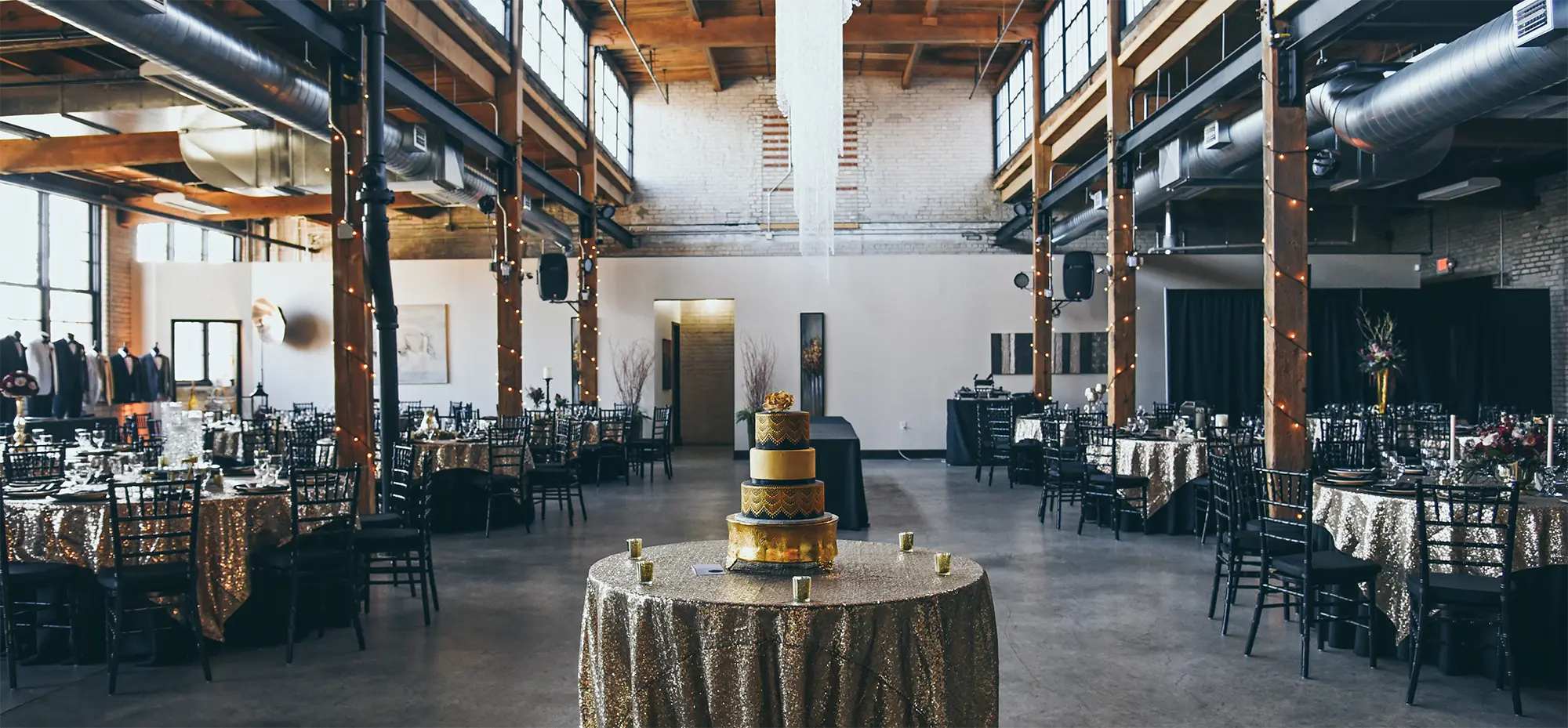 Interior landscape photograph of a large room area showing many tables and black chairs with gold colored glitter table covers over the tables and one of the tables in the front middle has a gold colored decorated four layer cake at the Studio D2D Event Space venue