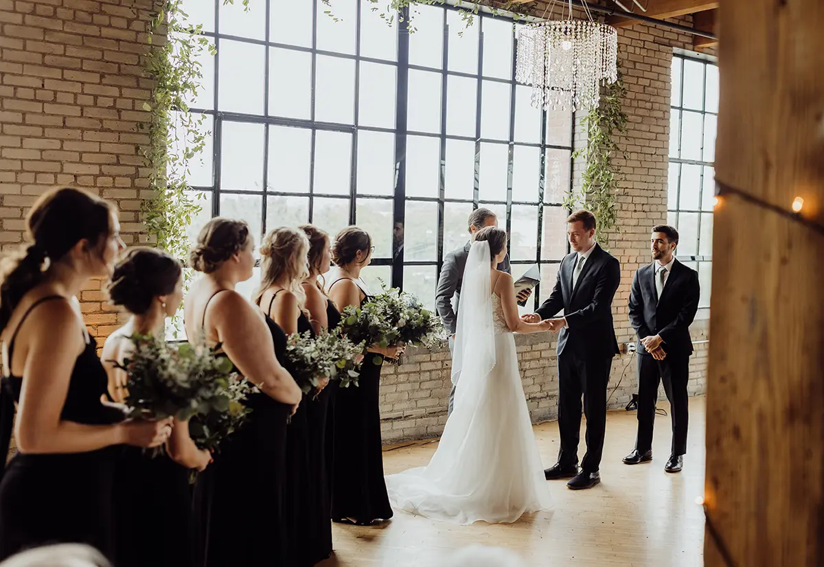 Close-up interior landscape photograph perspective of a wedding ceremony in session with the bride and groom holding hands as one of the groomsmen and six bridesmaids look at the new married couple at the Studio D2D Event Space venue