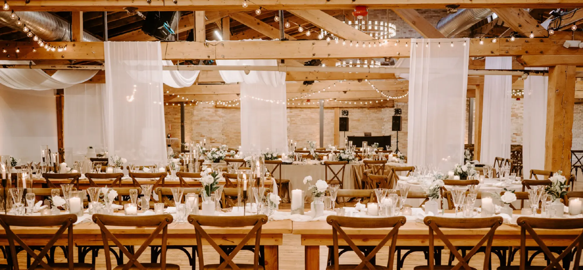 Large area filled with wooden tables and chairs, wedding drapes hung all over, and a DJ music setup station inside at The Goei Center venue