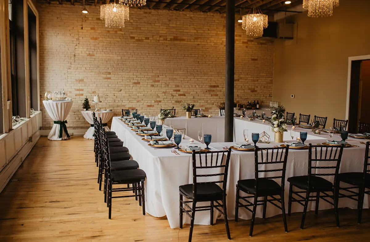 Long rectangular white table set up for a wedding party with black chairs all around, dark bronze colored plates, and dark blue colored glass cups as there are numerous crystal glass chandeliers floating above inside The Harris Building venue