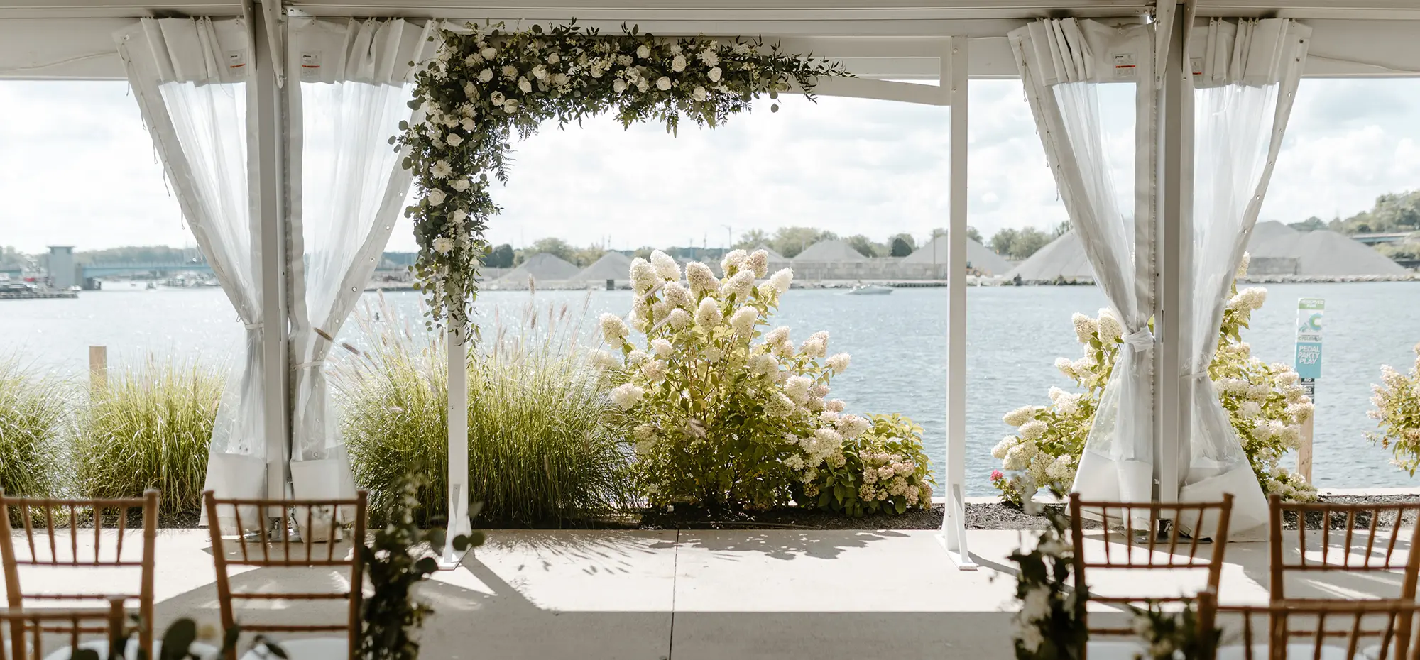 Outdoor landscape photograph of a white wedding overhead canopy display with a white flower arch decoration and a few chairs nearby as the setup is overlooking a body of water at The Inn at Harbor Shores venue
