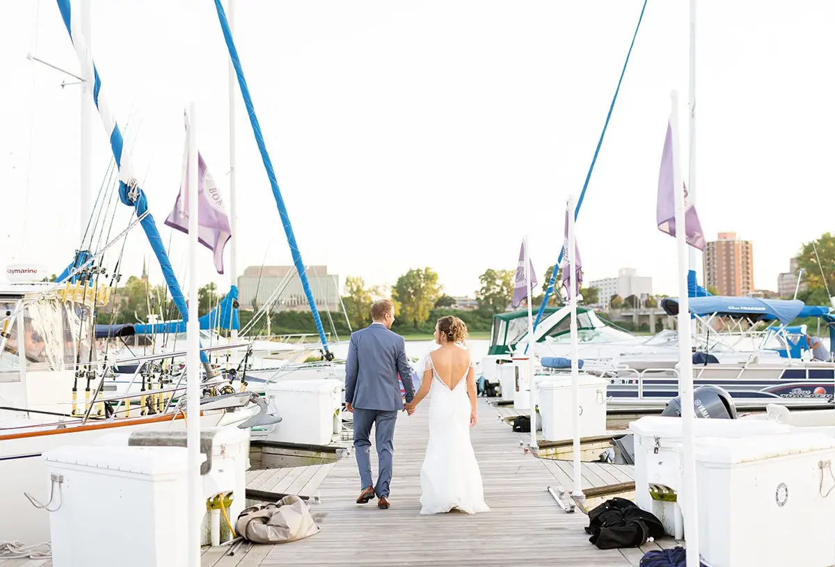 Bride and groom are holding hands while walking on a dock loading area with several motorboat vehicles around outside at The Inn at Harbor Shores venue