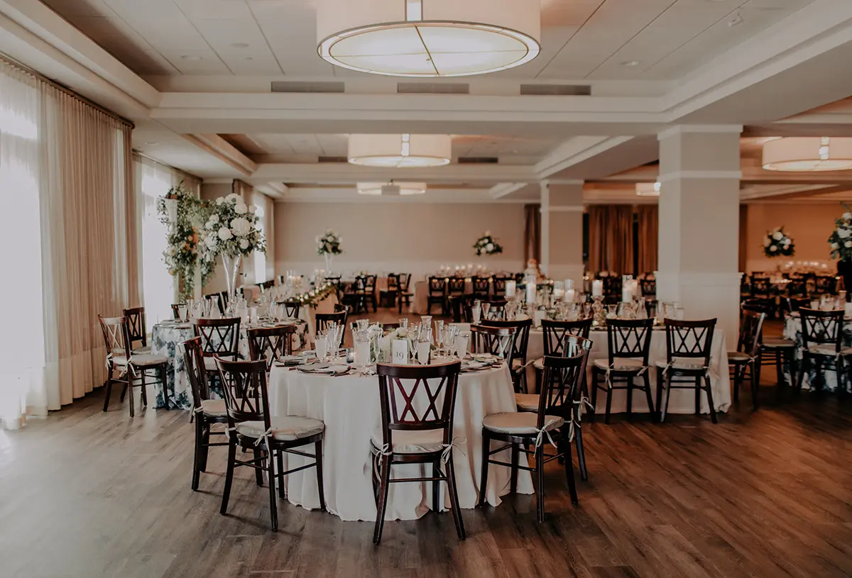 Indoor landscape photograph of several white tables spread out with dark bronze colored chairs and other flower bouquets spread out across the tables at The Inn at Harbor Shores venue