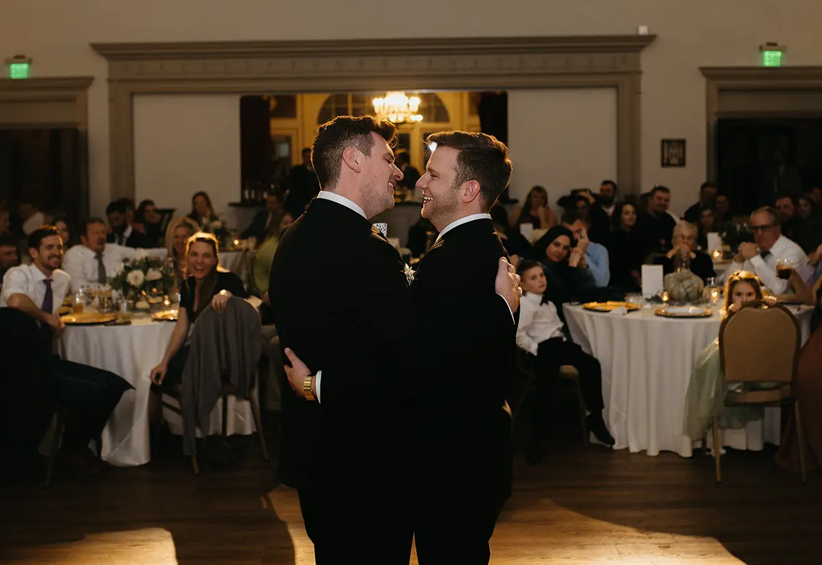 Two men in suits hugging in a ballroom area as they dance together and smile at each other while the audience glances upon them inside the Lit venue