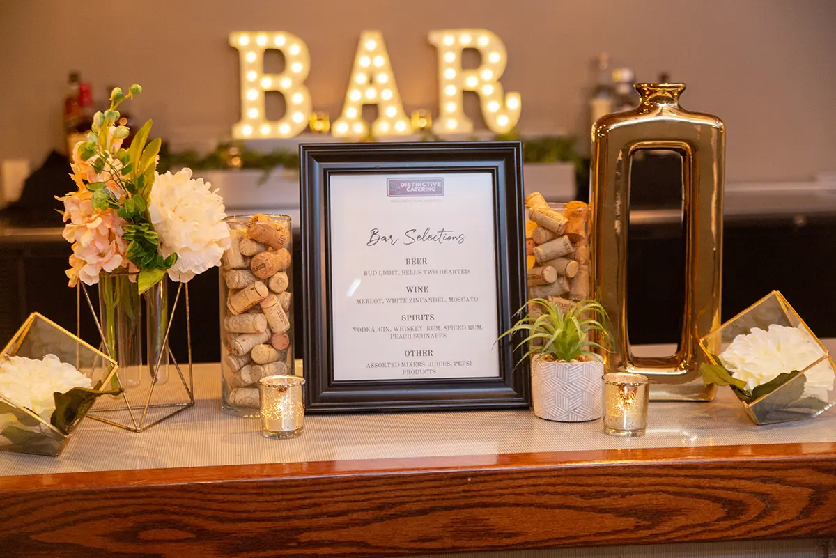 Close-up photograph perspective of a table with a picture frame titled "Distinctive Catering" and "Bar Selections" listings underneath in the foreground alongside other wedding decorative objects with a illuminated "BAR" sign in the background