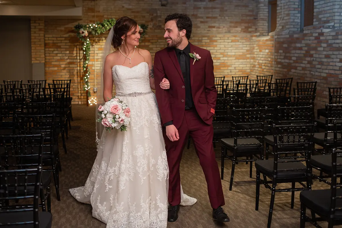 Bride and groom standing as they smile at each other in the middle aisle (groom is in a dark burgundy suit) inside The Waddell Center venue while there are other black empty chairs around them