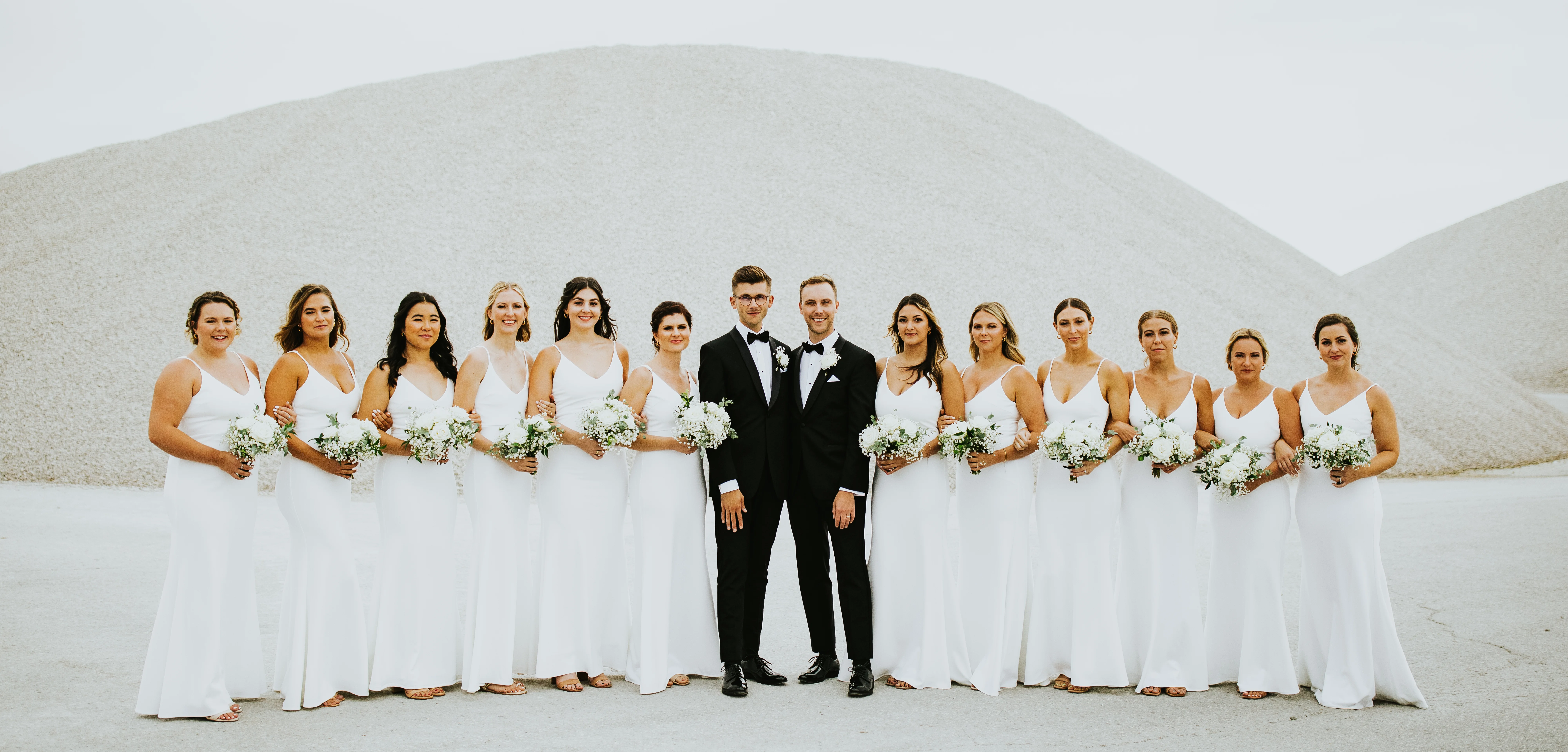 two grooms wearing black tuxes standing between their bridesmaids wearing white