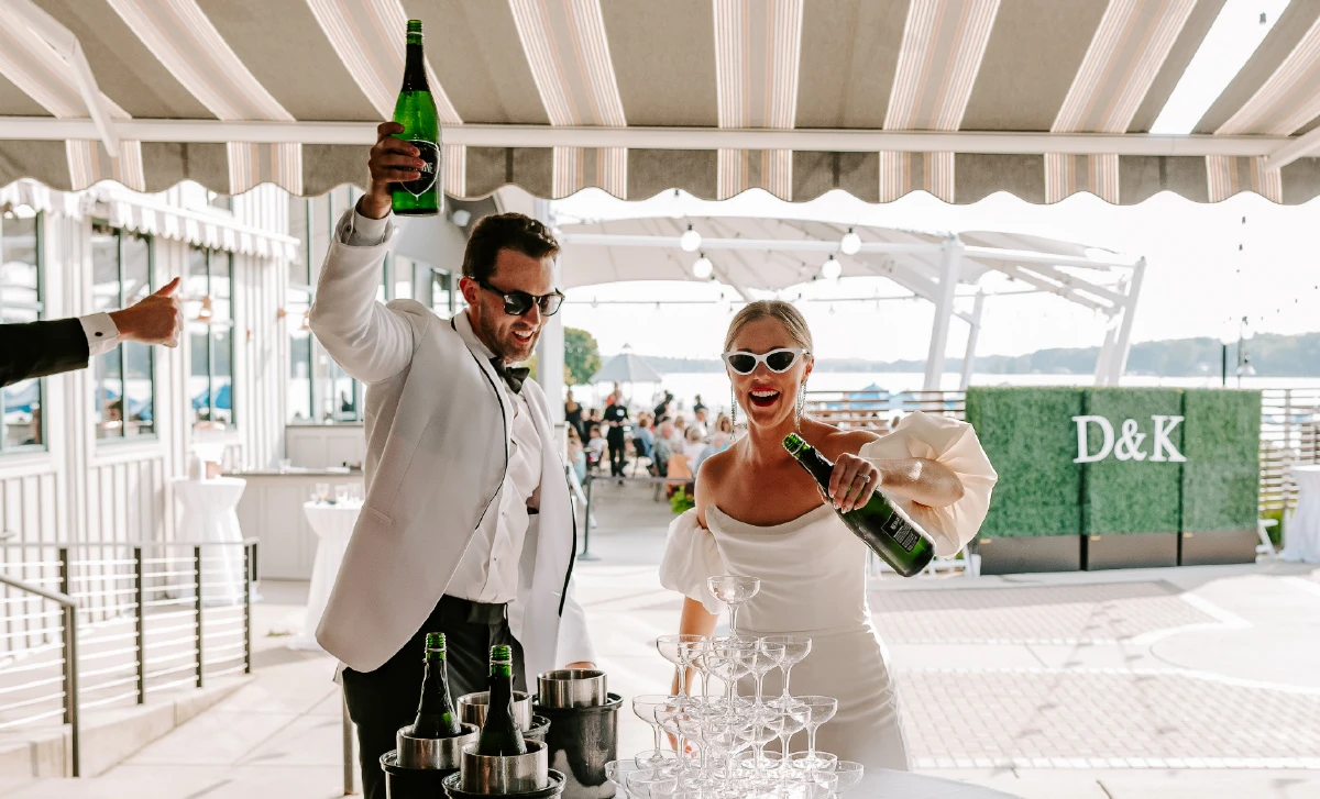 bride and groom pouring champagne on top of glass cup pyramid while wearing sunglasses