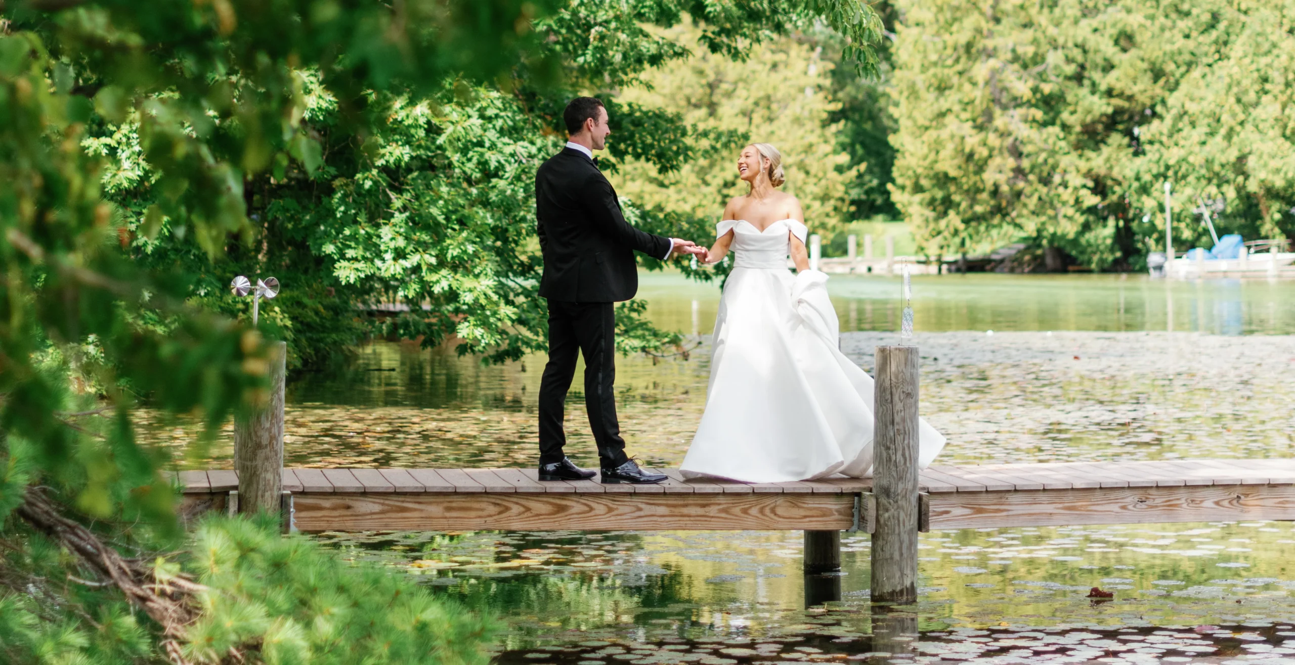 bride and groom holding hands on wooden dock on lake in wooded area