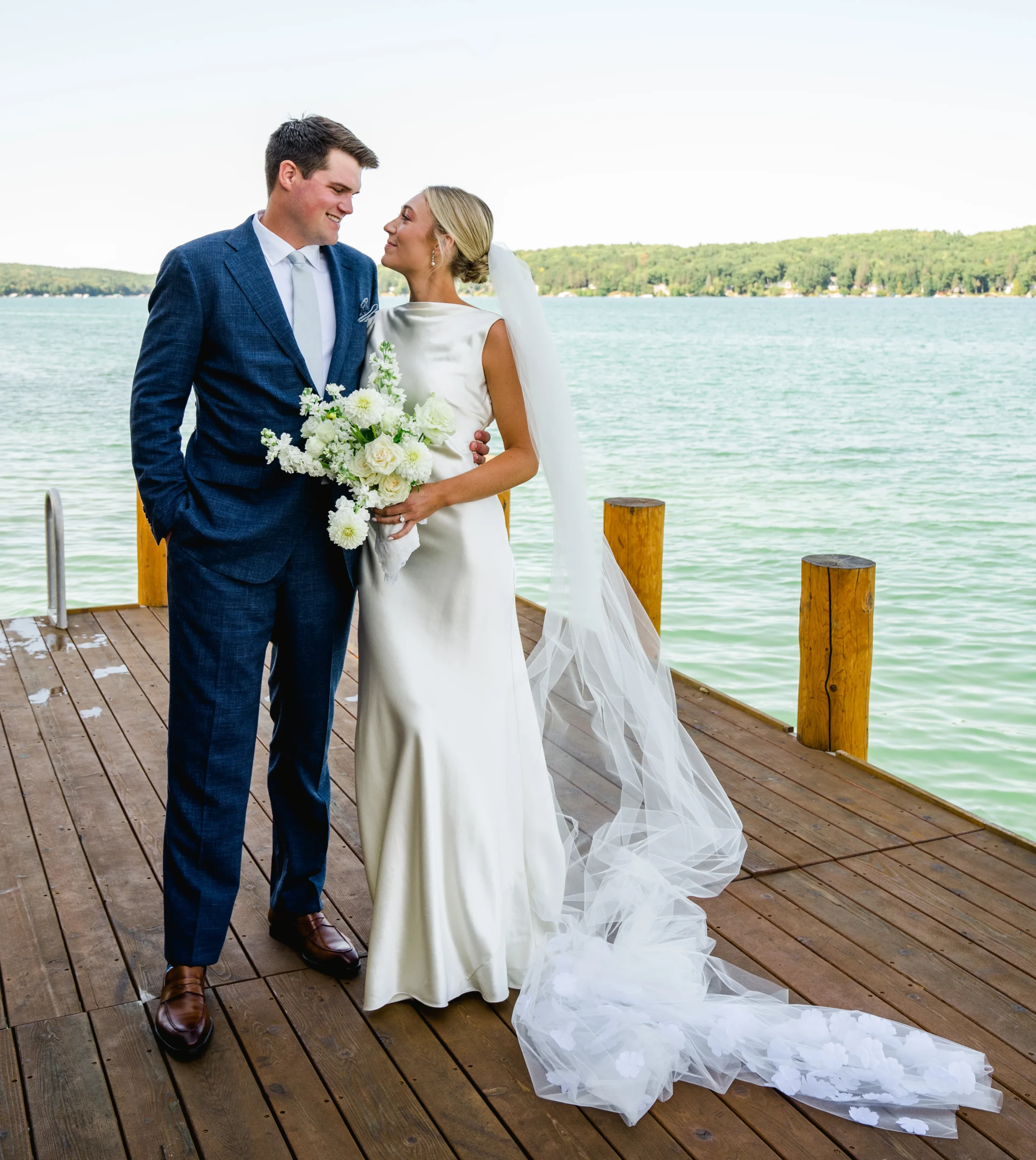 bride and groom looking at each other while standing on a dock in front of water