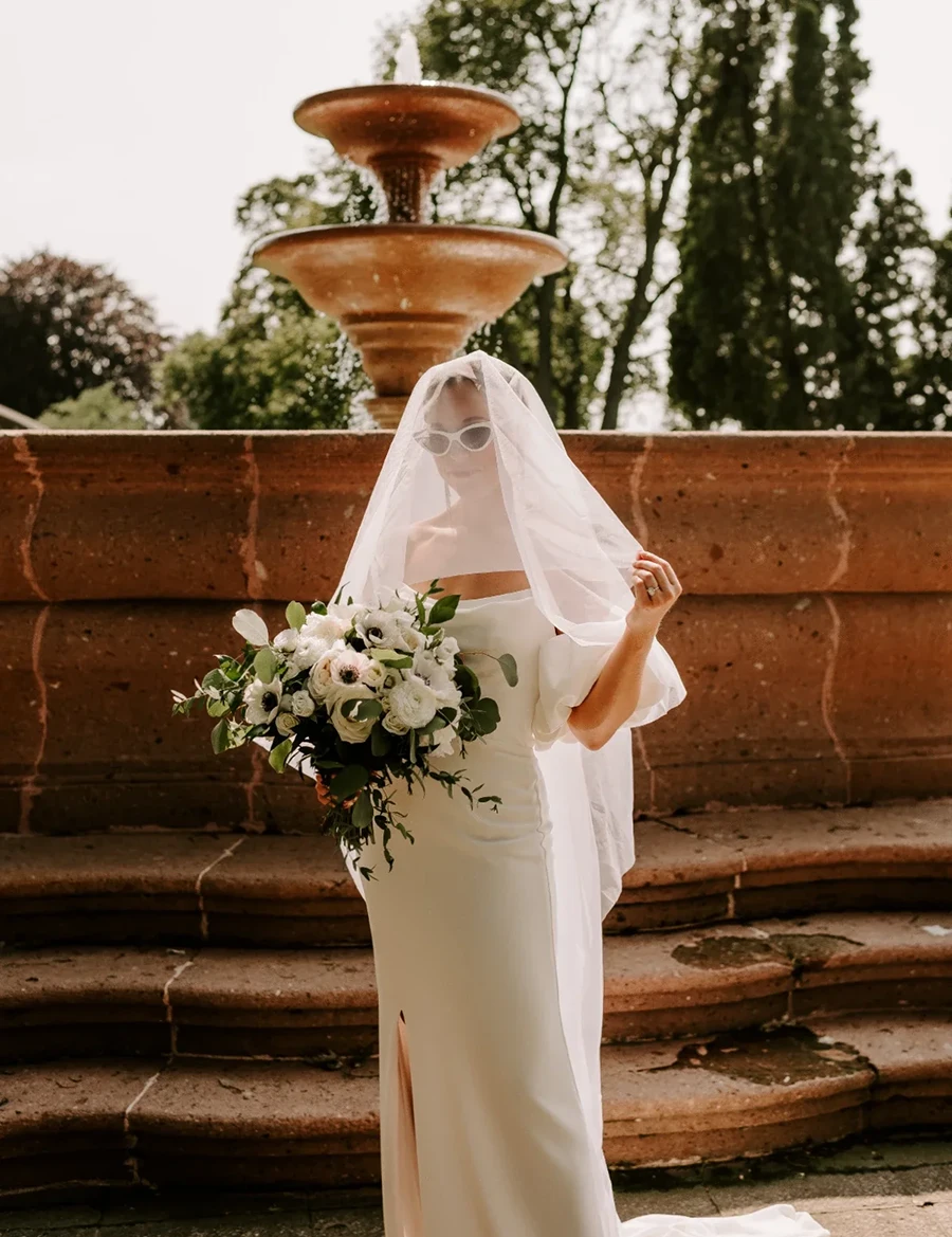 bride in front of fountain wearing cat eyed sunglass and veil covering her face